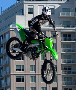 Reference photo for Jackalope Jump: a motocross rider on a green bike in mid-air in front of a building