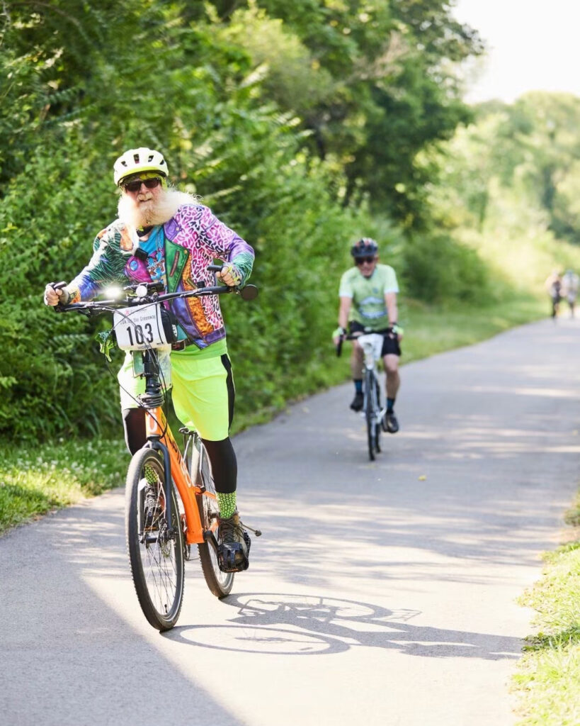 Reference photo for Light Up the World: a rider in a brightly colored outfit pedaling along a paved path with a second rider behind
