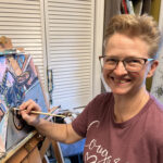 Kristi Gray smiling at her easel with a paintbrush in hand, working on “Wheels Up”