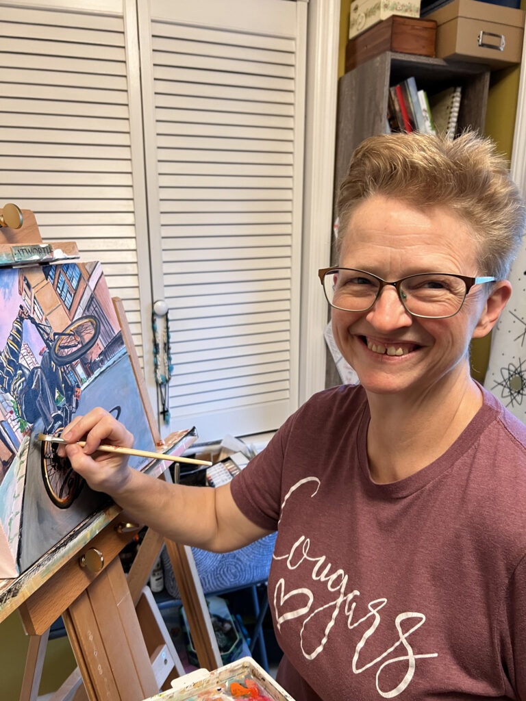 Kristi Gray smiling at her easel with a paintbrush in hand, working on “Wheels Up”