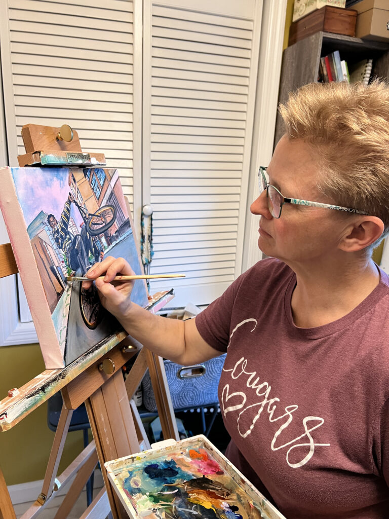 Kristi Gray painting a bicycle wheel in “Wheels Up”, with her palette in the foreground